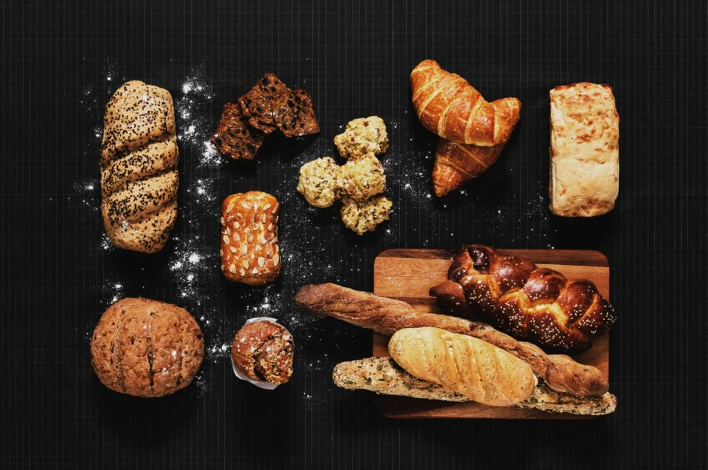 Assorted breads and pastries on a black background, including baguettes, croissants, and muffins, with flour sprinkled around, creating a rustic feel.