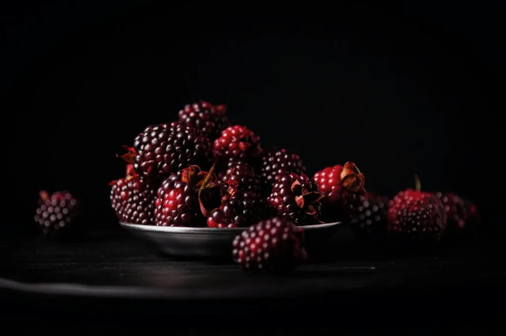 A plate of fresh, dark red blackberries is lit dramatically against a black background, emphasizing their rich color and glossy texture.
