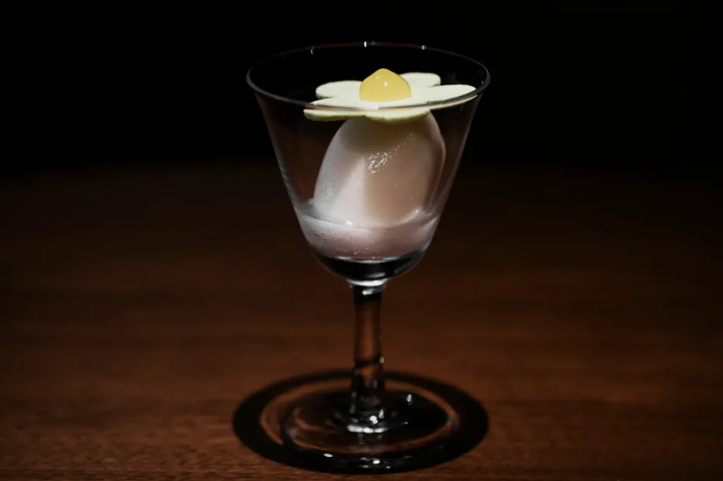 A delicate cocktail in a glass, with a pale flower-shaped garnish on top, featuring a small yellow drop at its center. The setting is elegant and dimly lit.