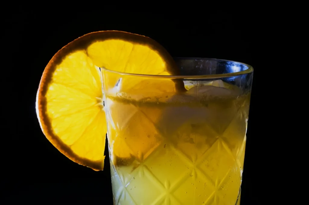 A textured glass filled with yellow liquid and ice against a black background, garnished with an orange slice, creating a refreshing and vibrant look.