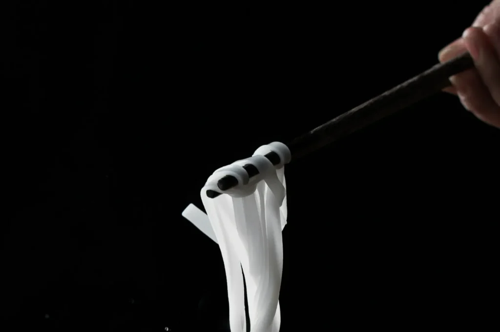 Chopsticks hold thin, glossy, white noodles against a dark background, emphasizing the noodles' texture and sheen. The mood is minimalist and elegant.