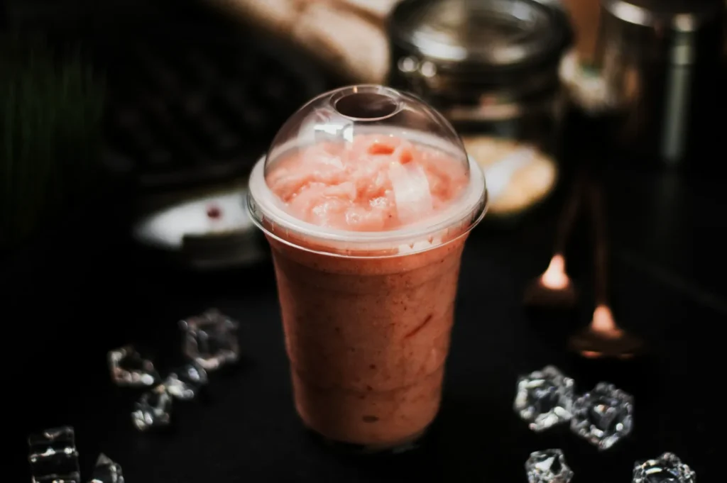 A pink smoothie in a clear plastic cup with a dome lid sits on a dark table. Nearby are ice cubes and blurred kitchen items, creating a cozy vibe.