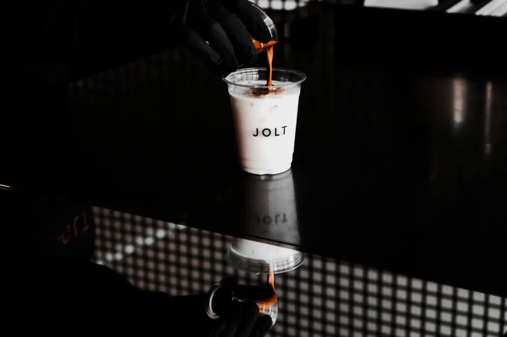 A gloved hand pours espresso into a plastic cup of iced milk labeled "JOLT" on a dark reflective surface, creating a moody and sleek atmosphere.