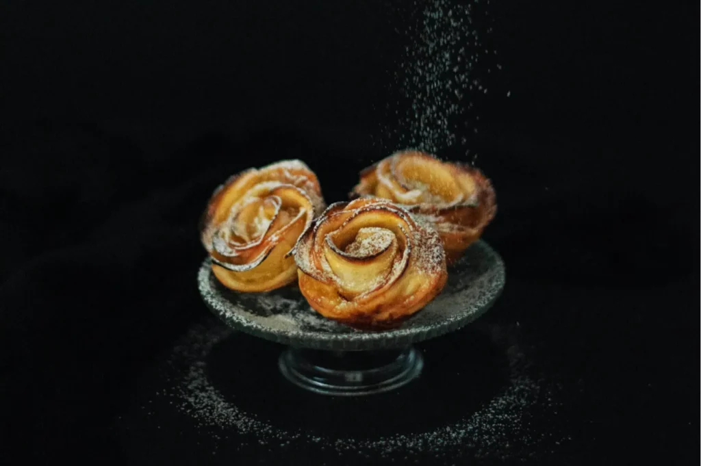 Three rose-shaped pastries on a pedestal, sprinkled with powdered sugar, against a dark background, evoking an elegant and tempting mood.