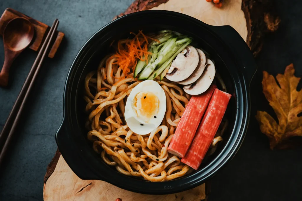 A black bowl with Asian noodles, topped with a halved boiled egg, crab sticks, sliced mushrooms, cucumber, and grated carrots, on a wooden board.