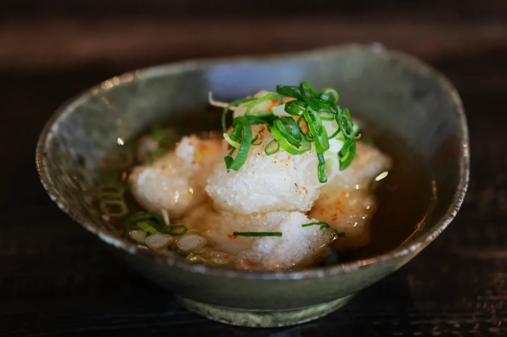 Bowl of soup with light-colored dumplings, garnished with sliced green onions. The bowl is rustic and set against a dark wood surface.