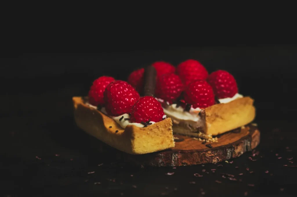 A square tart topped with vibrant red raspberries and a chocolate stick rests on a rustic wooden slab. The mood is rustic and inviting.