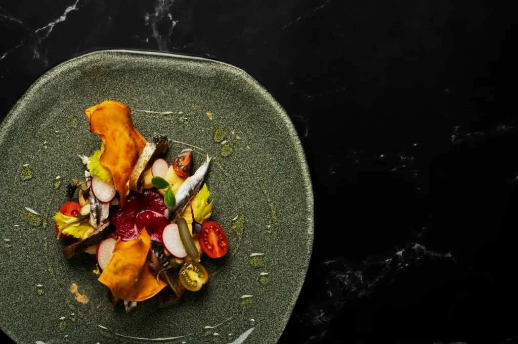 A beautifully plated dish on a textured green plate with vibrant veggies, thin fish fillets, and crispy chips, evoking a sophisticated gourmet feel.