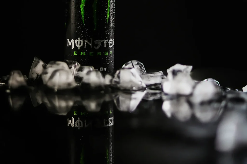A can of Monster Energy stands upright on a glossy black surface surrounded by scattered ice cubes, conveying a cold, refreshing vibe.