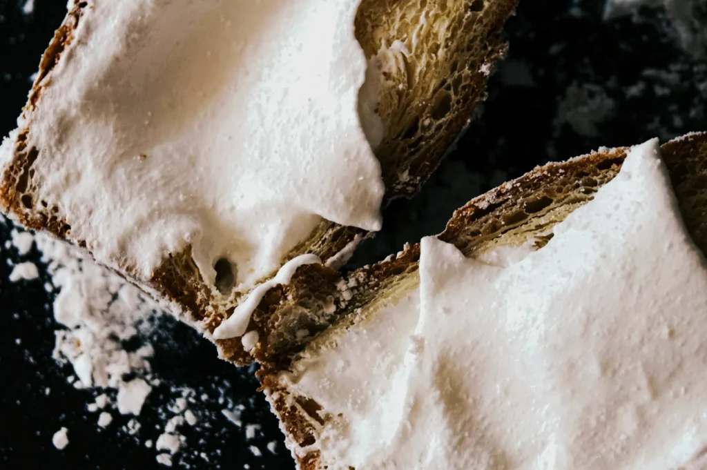 Close-up of two pieces of toast with thick, creamy spread on a dark surface dusted with powdered sugar, conveying a cozy, indulgent feeling.