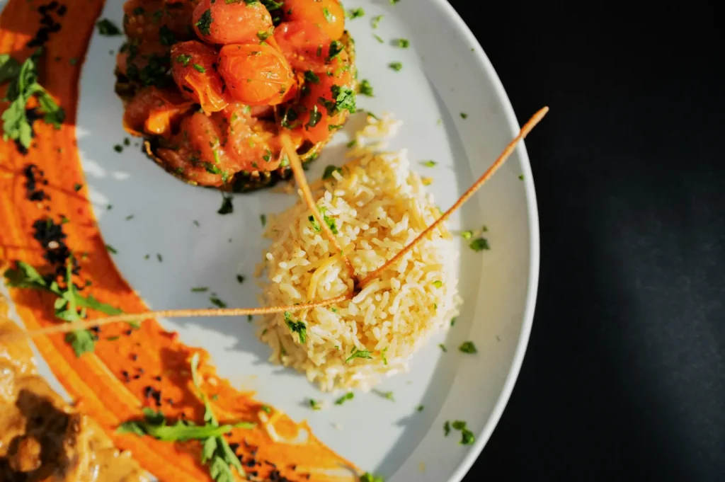A white plate with rice garnished with herbs, roasted cherry tomatoes, and a vibrant red sauce swirl. The dish looks flavorful and artfully arranged.