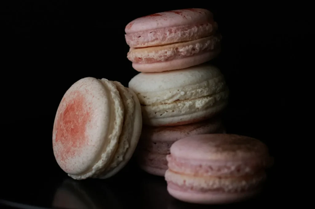 A pile of assorted pink and white macarons with a dusting of cocoa powder. The dark background highlights their delicate texture and pastel colors.