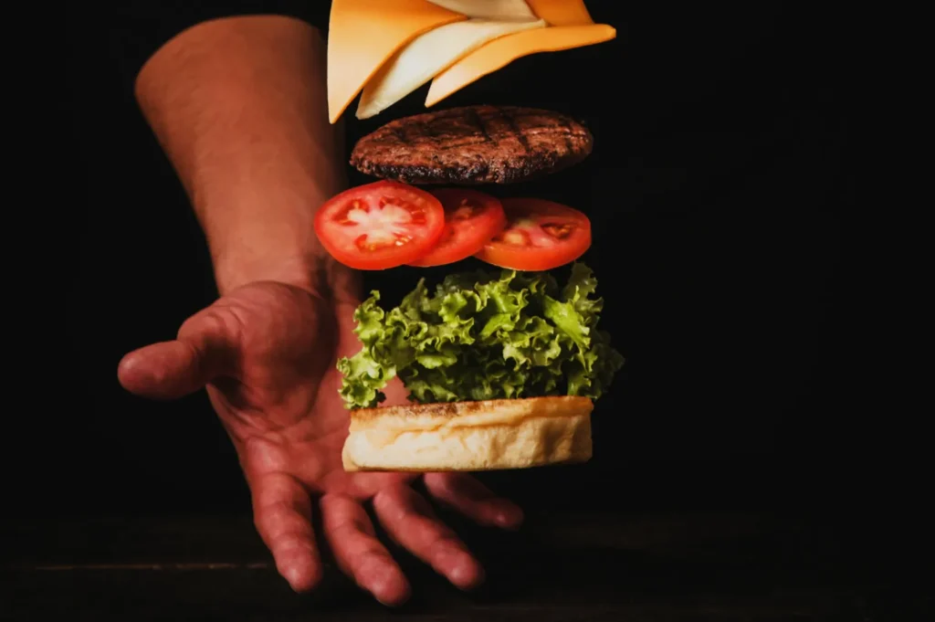 Deconstructed burger floating with layers of cheese, patty, tomato, lettuce, and bun against a dark background and a person's outstretched hand.