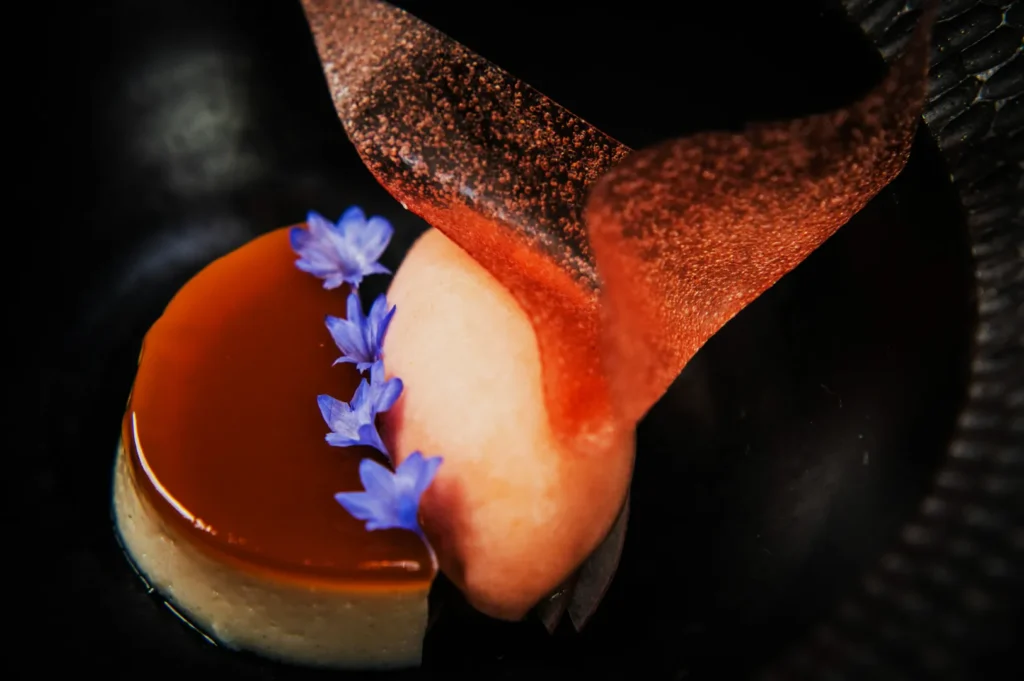 Elegant dessert with caramel-topped flan, adorned with delicate purple flowers and a curved chocolate shard on a dark plate. Sophisticated and artistic.