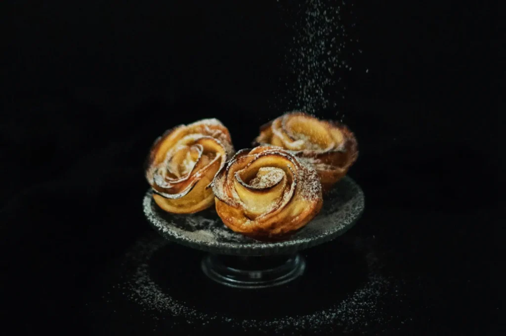 Three apple rose pastries dusted with powdered sugar sit elegantly on a glass stand against a black background, creating a delicate and appetizing display.