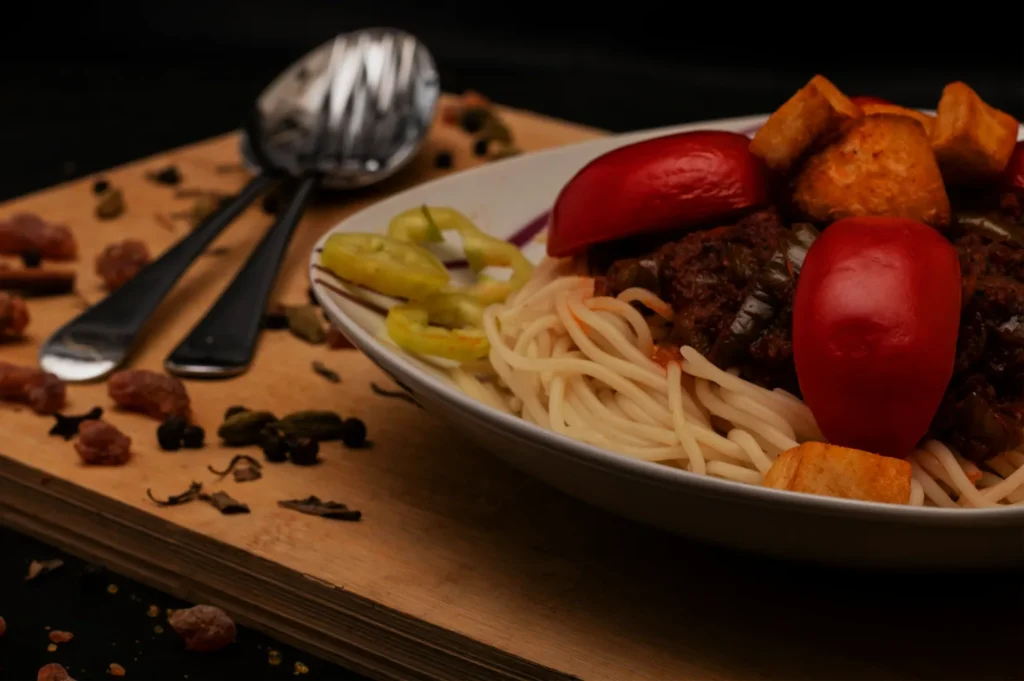 A bowl of spaghetti topped with vibrant red bell peppers, golden fried tofu, and rich sauce, garnished with sliced green peppers. Cutlery and spices beside.