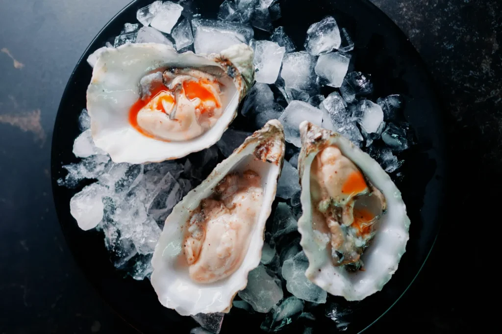 Three fresh oysters on a black plate with crushed ice. The oysters have a vibrant orange sauce, contrasting with the dark background, evoking freshness.