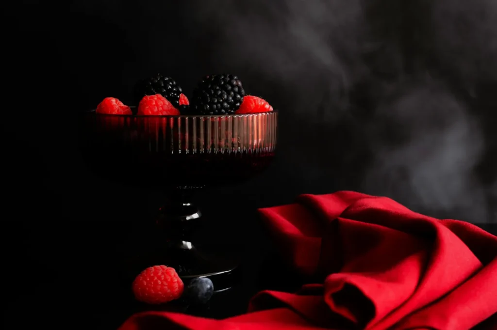 A dark glass dish holds blackberries and raspberries, surrounded by a red cloth on a black surface. The backdrop is a smoky, matte black.