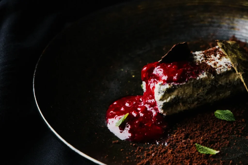 Dark plate with a slice of creamy dessert topped with rich red berry sauce. Cocoa powder and mint leaves add a touch of elegance.