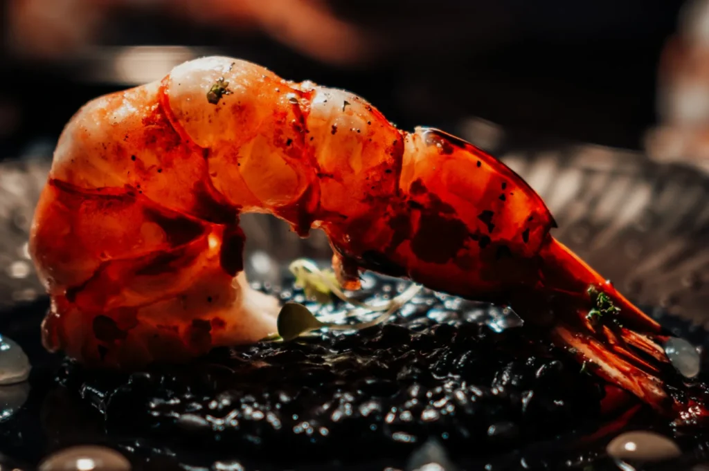 Close-up of a grilled prawn with a glistening orange shell, artistically placed over black rice. The setting is elegant, warm, and inviting.