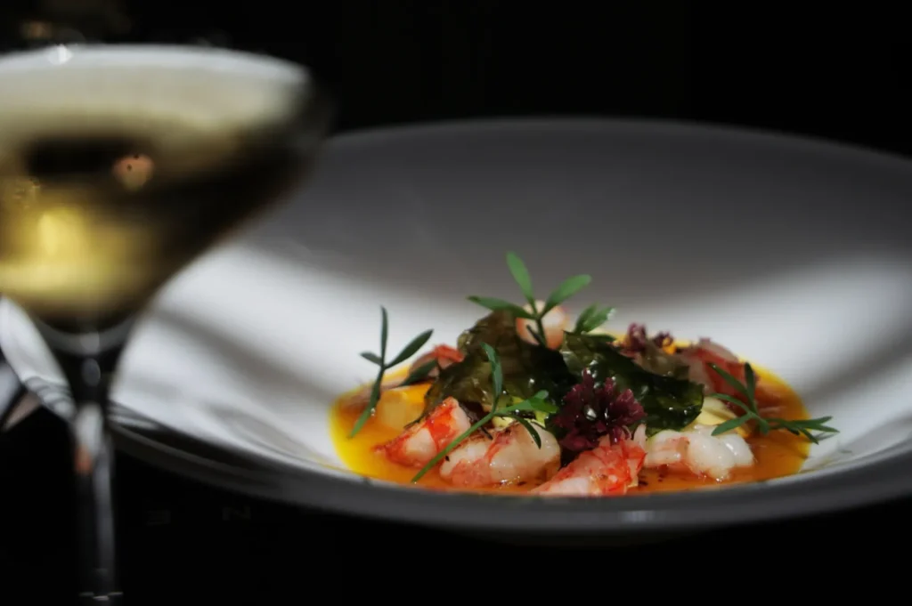 A gourmet dish of vibrant prawns in yellow sauce with green garnish and purple flowers on a white plate. A glass of white wine is blurred in the foreground.