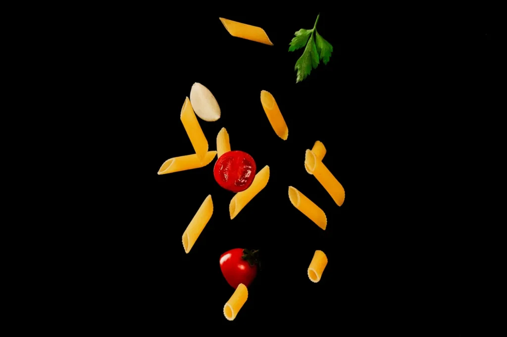 Penne pasta, cherry tomatoes, a garlic clove, and parsley float against a black background, creating a dynamic and vibrant culinary scene.