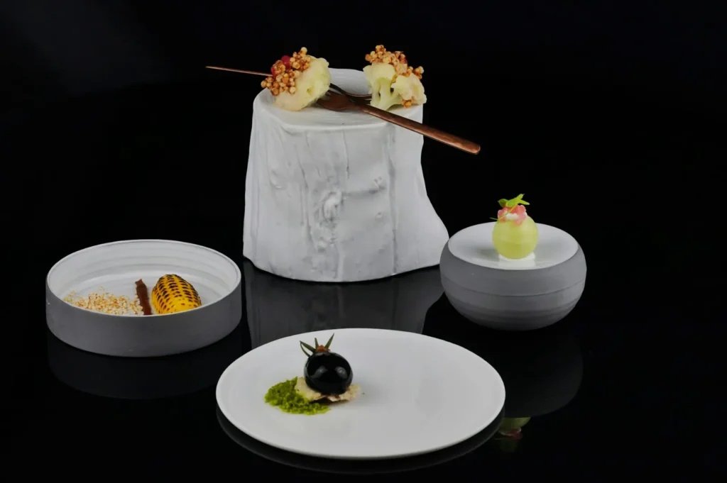 Elegant gourmet dishes on a black reflective surface: spherical green dessert, grilled corn in a dish, black mousse with herb garnish, and cauliflower blooms with crunchy toppings.