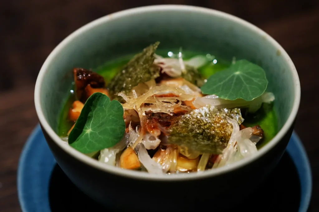 A vibrant dish in a blue bowl, featuring green sauce, leafy greens, translucent toppings, and crunchy elements, exuding a fresh, gourmet appeal.