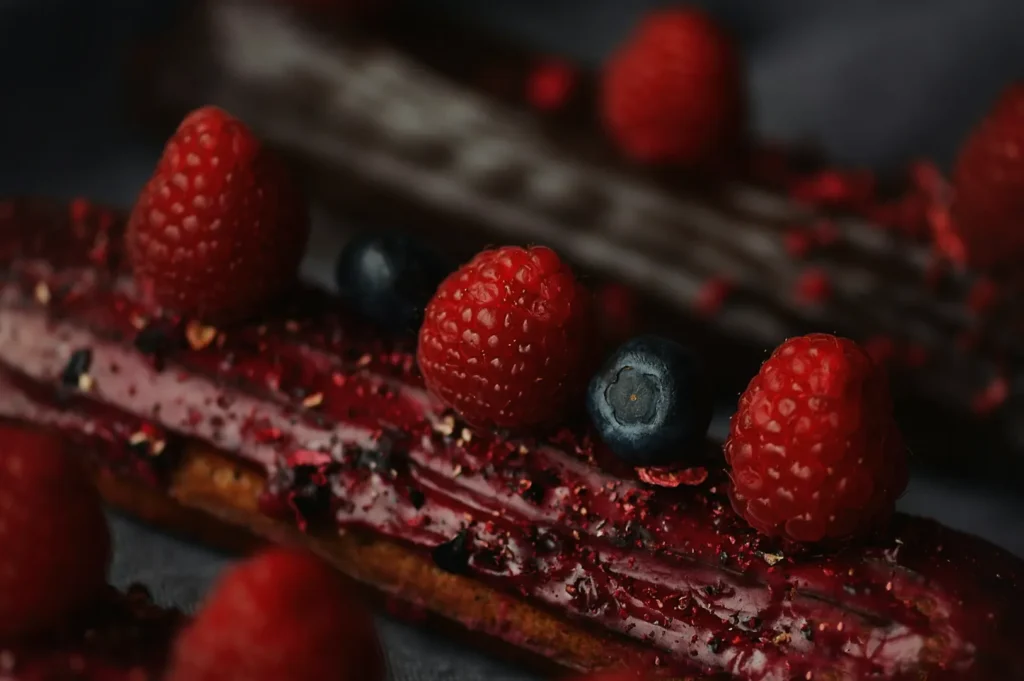 Close-up of glazed eclairs topped with fresh raspberries and blueberries. The rich red and deep blue colors contrast against a dark background, conveying a gourmet feel.