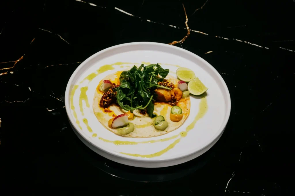 A gourmet taco on a white plate with grilled toppings, leafy greens, radish slices, and lime wedges, arranged artistically on a black marble surface.