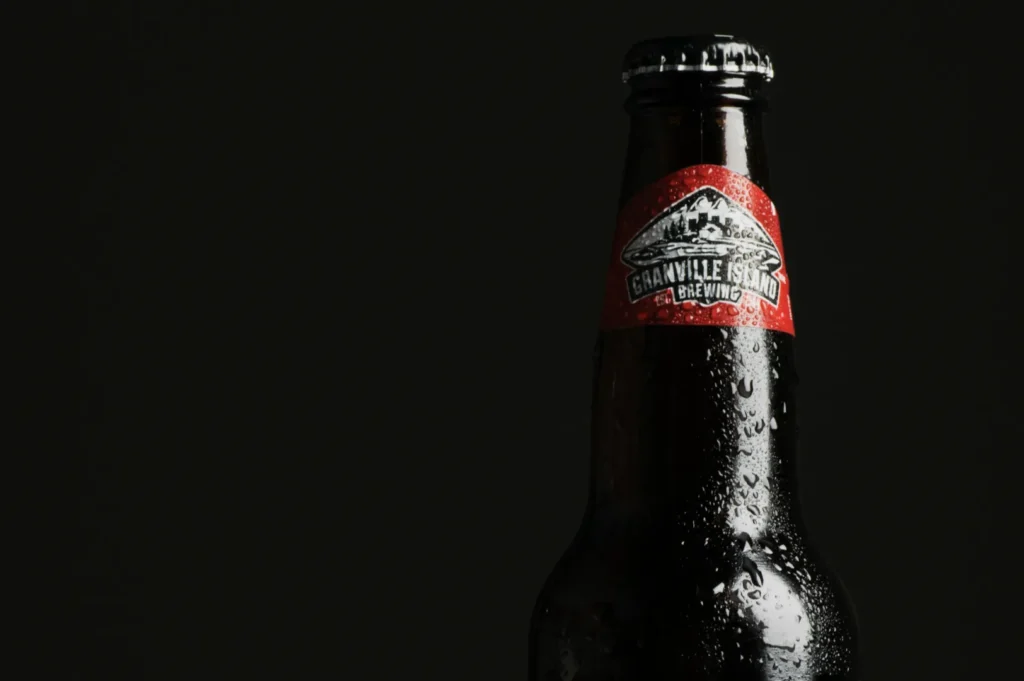 Close-up of a dark beer bottle with a red and silver label showing "Granville Island Brewing." Water droplets bead on the bottle, set against a dark background, conveying refreshment.