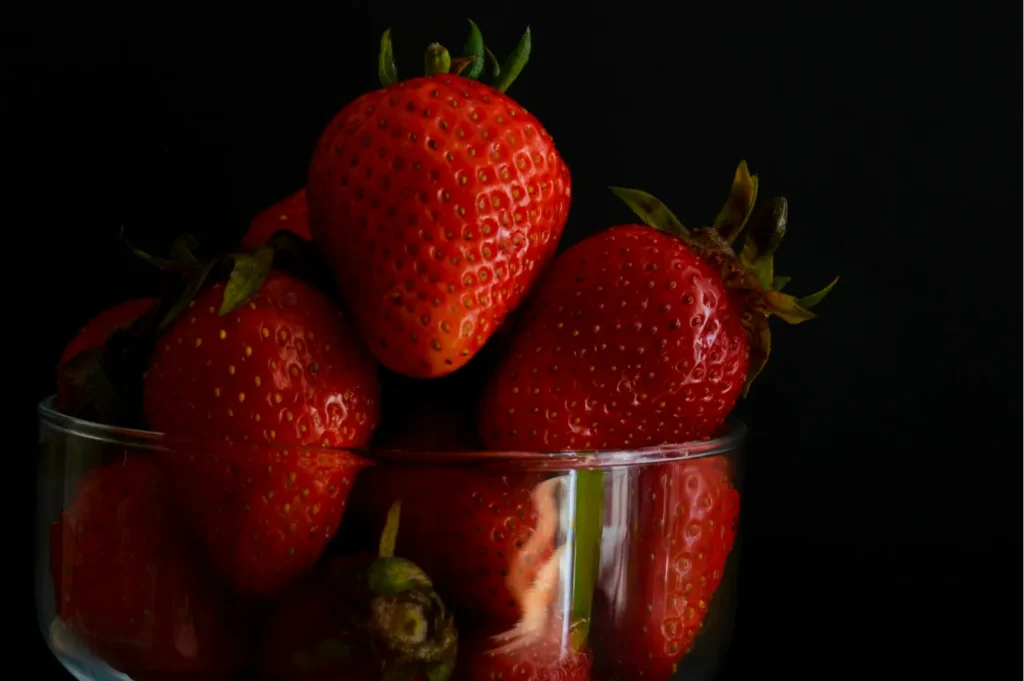 A glass bowl filled with vibrant red strawberries, accented by their green leaves. The dark background highlights the fruit's fresh, juicy appearance.