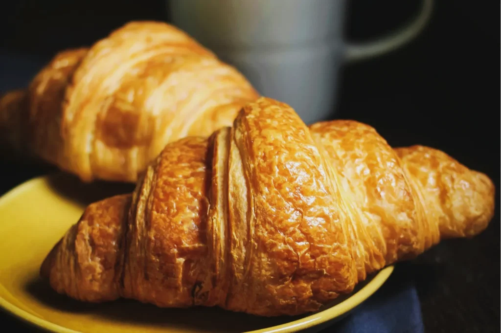 Two golden-brown croissants on a yellow plate with a blurred mug in the background, conveying a cozy breakfast scene. Soft lighting highlights the flaky texture.
