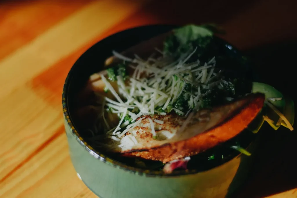 A bowl of salad on a wooden table, topped with grated cheese and toasted bread. Natural light highlights fresh greens and adds a warm, rustic feel.