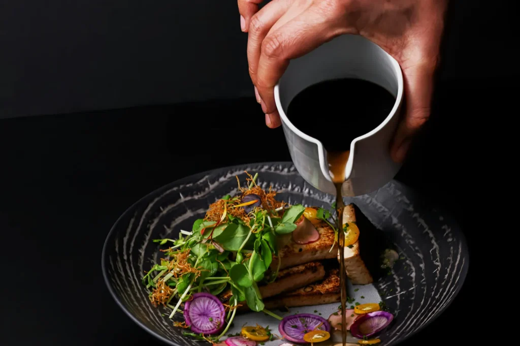 A hand pours dark sauce from a white pitcher onto a gourmet dish of grilled tofu, microgreens, and radish slices on a black plate, creating an elegant scene.