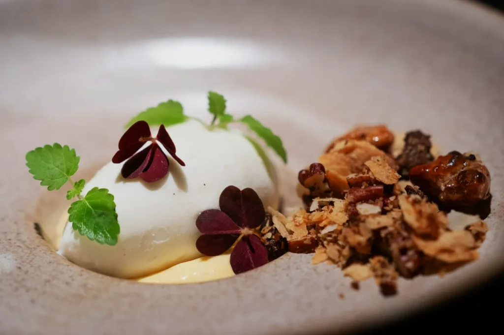 Elegant dessert featuring a scoop of creamy mousse, garnished with green and red leaves, beside a cluster of caramelized nuts and crispy flakes on a ceramic plate.