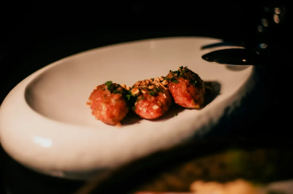 Three small meatballs garnished with herbs and nuts on a white, oval plate. The dim lighting creates a warm, inviting atmosphere.