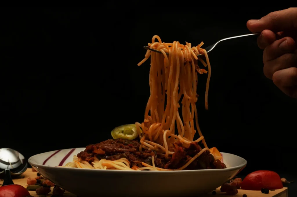 A hand holds a fork lifting spaghetti with meat sauce from a bowl. A slice of green pepper is visible. The background is dark, highlighting the dish.