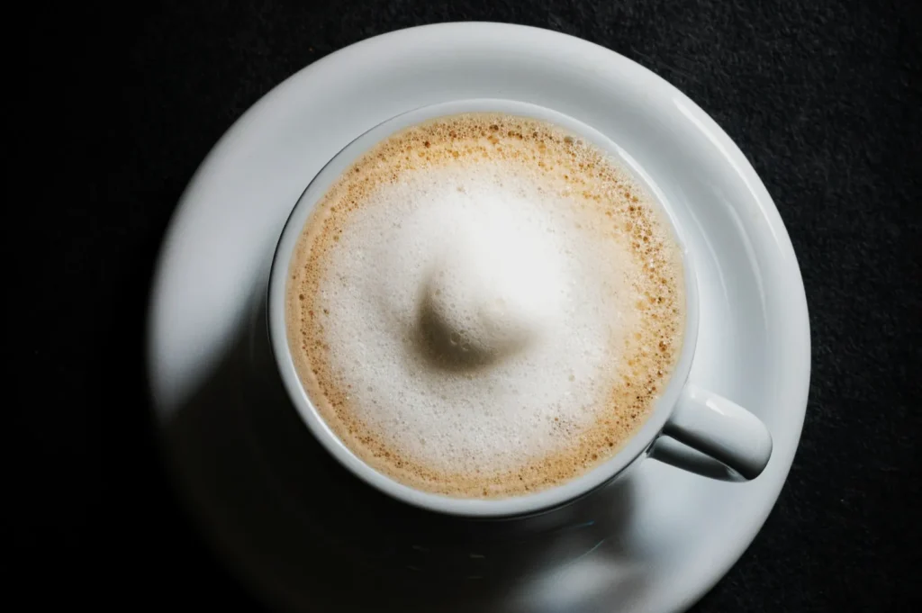 Top view of a frothy cappuccino in a white cup on a saucer, set against a dark background. The foamed milk creates a soft, inviting texture.