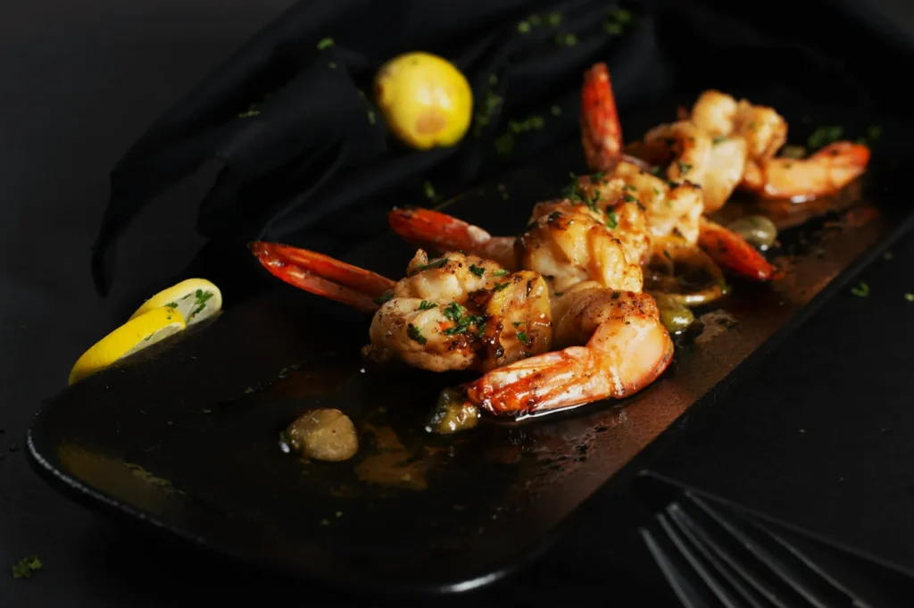 Grilled shrimp skewers garnished with herbs on a black rectangular plate. Background includes a lemon and a black cloth, creating an elegant presentation.