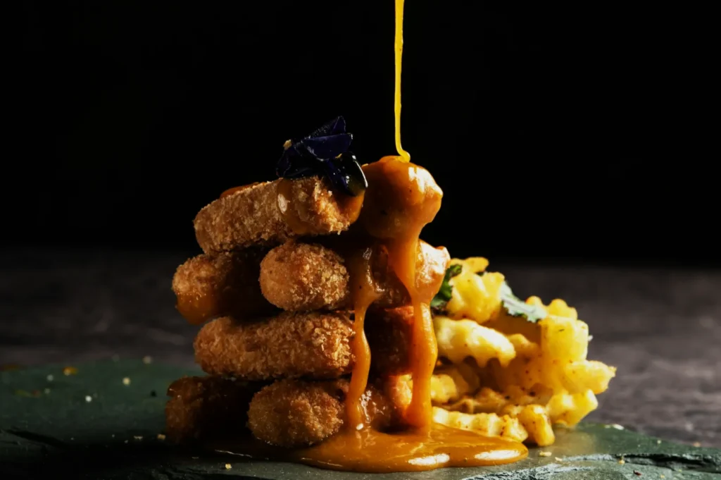 Golden sauce drips onto a stacked tower of breaded croquettes, garnished with a purple flower, with crinkle-cut fries on the side, set against a dark background.
