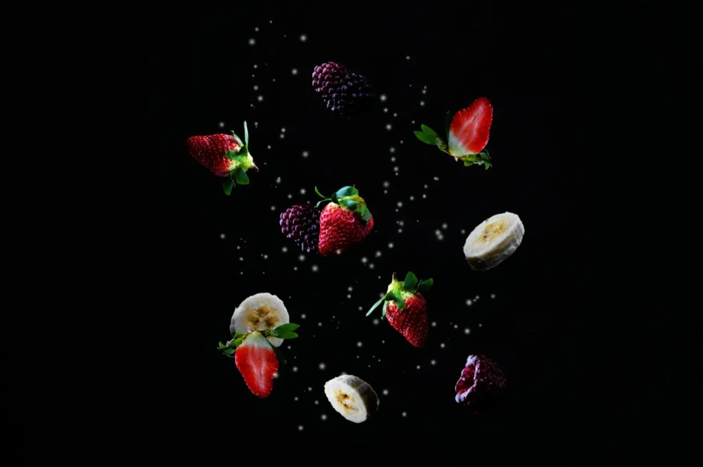 Assorted fruits, including strawberries, blackberries, raspberries, and banana slices, artistically suspended in mid-air against a dark background.