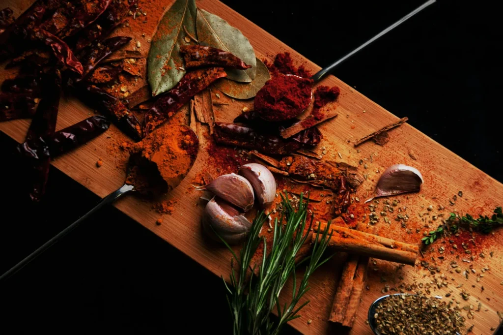 A wooden board with an assortment of spices including chili powder, bay leaves, garlic, rosemary, cinnamon sticks, and dried chilies. Rustic and aromatic scene.