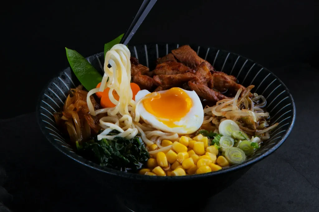 A black bowl of ramen with noodles, soft-boiled egg, corn, greens, sliced meat, snow peas, carrots, and spring onions. Chopsticks lift noodles, creating a savory, appetizing look.