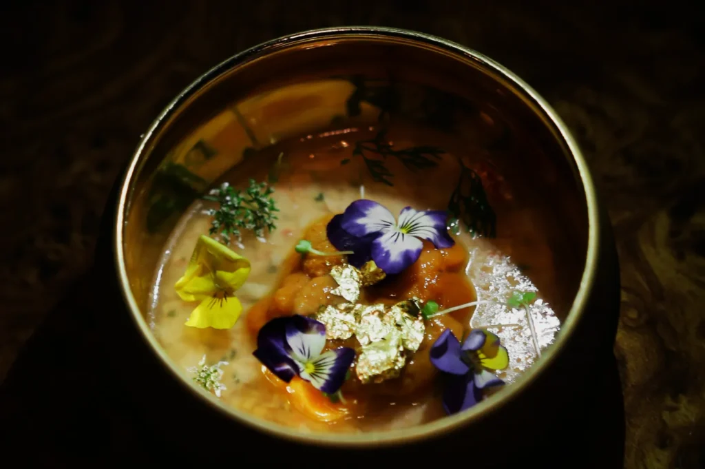 A luxurious dish in a golden bowl featuring uni, adorned with edible flowers and gold leaf. The presentation is elegant and artistic.