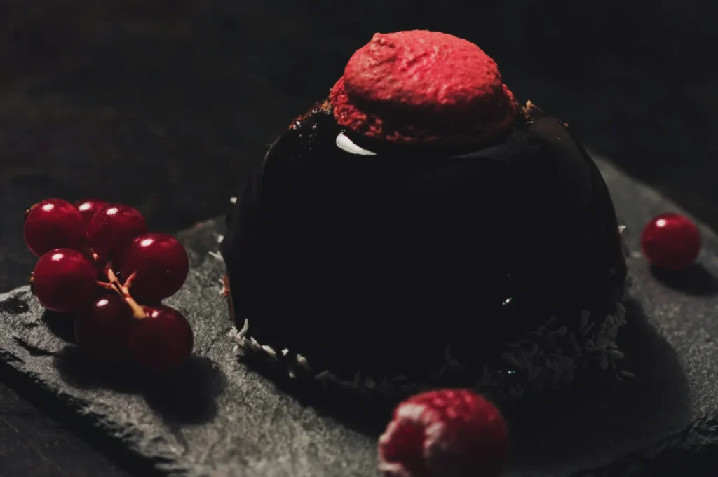 A glossy black dessert topped with a red macaron sits on a dark slate surface. Red currants and a raspberry surround the treat, creating a rich, elegant tone.