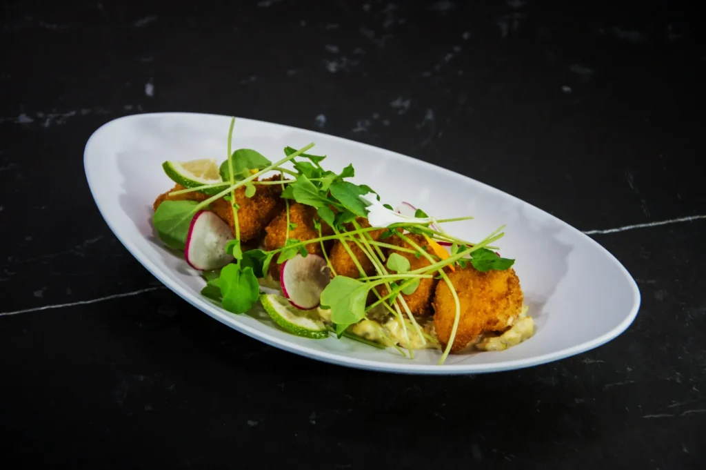 Four golden-brown croquettes on a white plate, garnished with fresh green arugula, radish slices, and lime wedges, atop a creamy sauce.