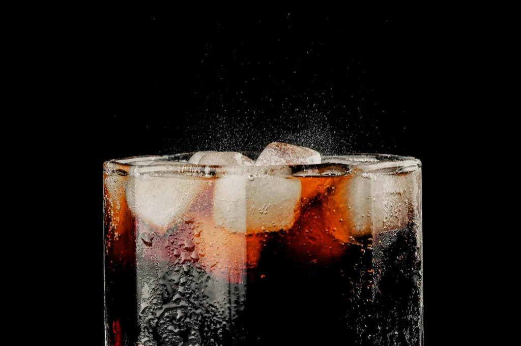 A glass of cola with ice cubes against a black background. Tiny bubbles and droplets on the glass add a refreshing, fizzy feel.