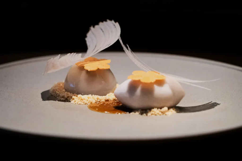 Elegant dessert with two white domes topped with orange flowers and delicate white feathers, resting on beige crumbs and caramel on a gray plate.