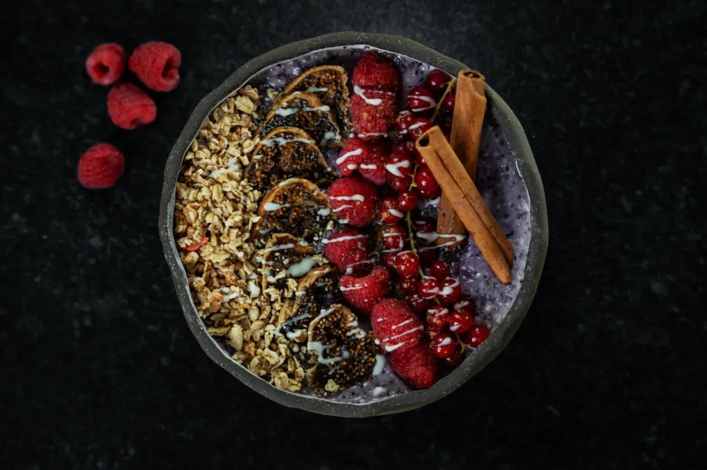 A vibrant smoothie bowl with blueberries, granola, figs, raspberries, and red currants, drizzled with white sauce and garnished with cinnamon sticks.